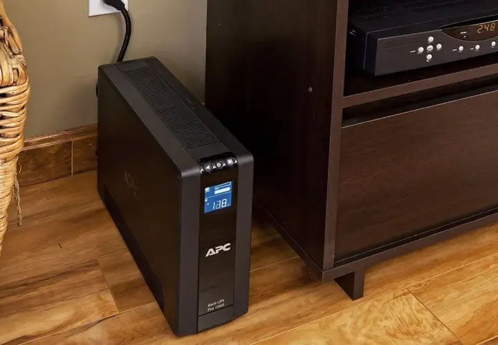 Ups co to? Czarny UPS APC Back-UPS Pro 1000 stoi na drewnianej podłodze obok mebla.