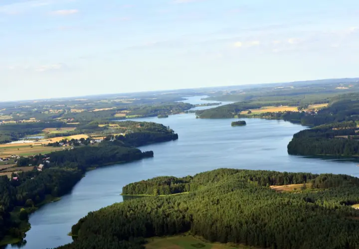 Malowniczy widok na jezioro Raduńskie Dolne otoczone lasami i polami. W oddali widać zabudowania.