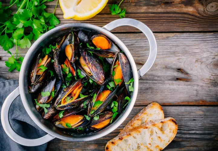 Un pentolino pieno di cozze alla marinara, guarnite con prezzemolo fresco e accompagnate da fette di pane tostato e spicchi di limone.
