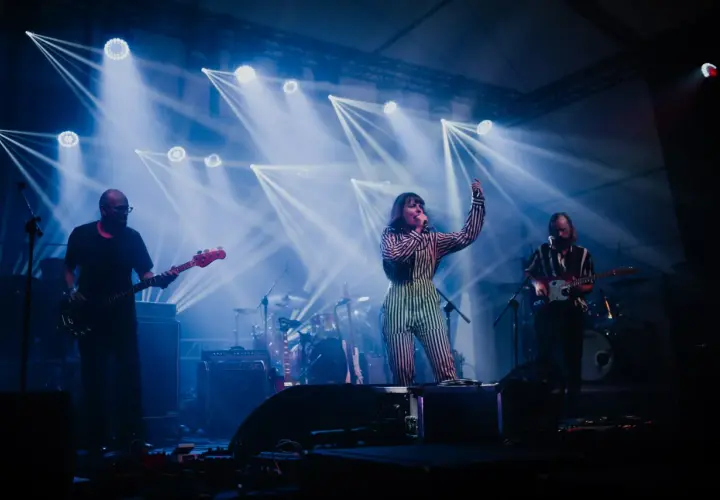 Izzy i The Black Trees na scenie, otoczeni niebieskimi światłami. Wokalistka w pasiastym stroju śpiewa, gitarzyści grają.