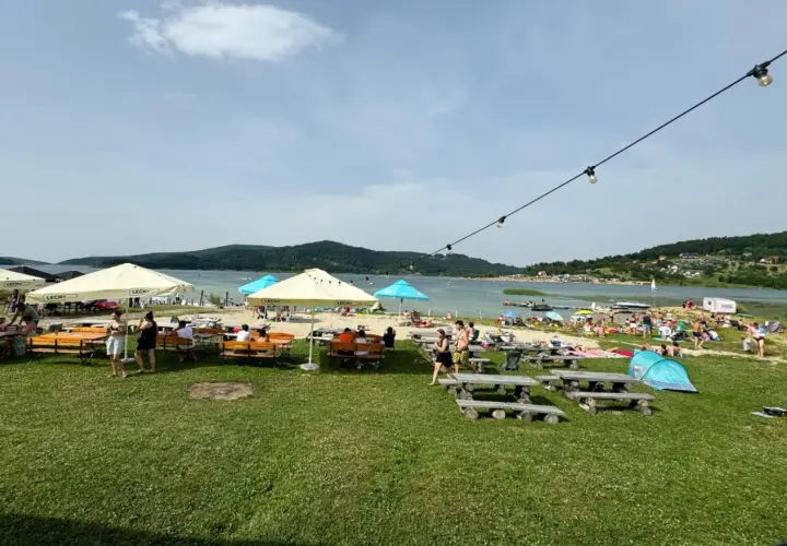 Słoneczny dzień nad Jeziorem Mucharskim. Ludzie odpoczywają na plaży, pod parasolami, przy stolikach. W tle jezioro i wzgórza. Atrakcje dla każdego.