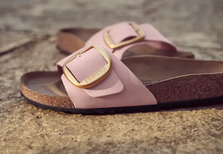Różowe klapki Birkenstock z złotą klamrą, leżące na kamiennym podłożu.