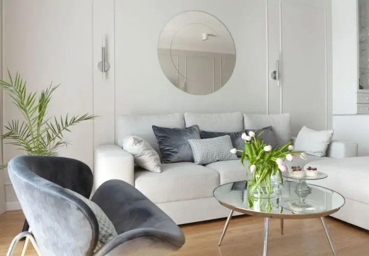 Przytulny salon z sofą i fotelami na tle jasnych, **białe ściany** zdobionych sztukaterią. Okrągłe lustro i bukiet tulipanów dodają elegancji.