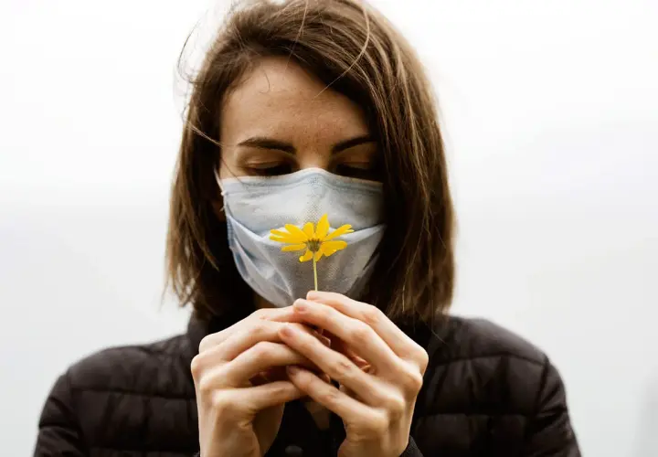 Mujer con mascarilla azul, oliendo una flor amarilla. Diversos tipos de mascarillas protegen, pero la esperanza florece.