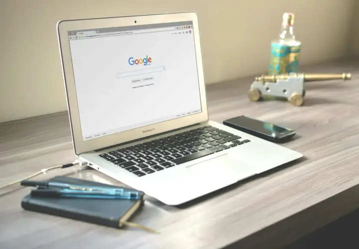 Laptop z otwartą stroną wyszukiwarki Google. Obok leży smartfon, notatnik z długopisami i ozdobne działko.
