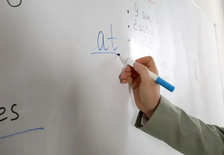 Mano scrive "at" su lavagna, spiegando il suo significato in inglese.
