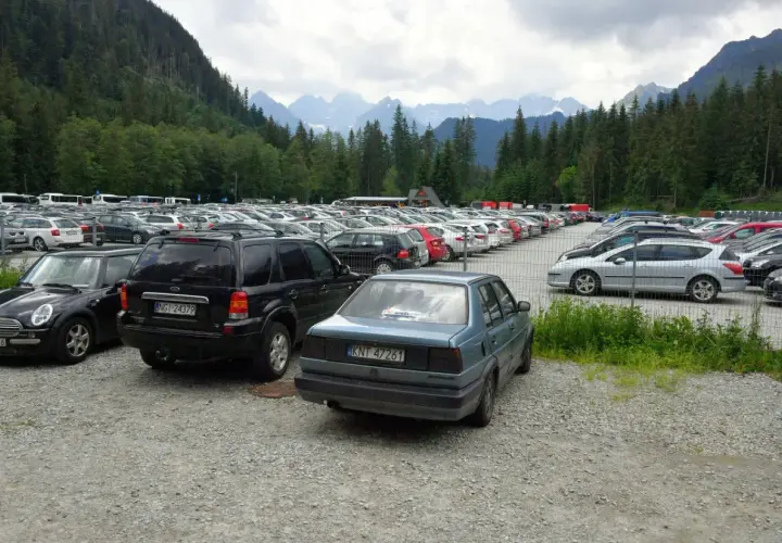 Duży parking samochodowy u podnóża gór, z widokiem na Tatry, jakby eic morskie oko patrzyło z góry.