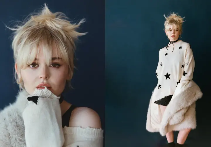 Emily Alyn Lind med rufsigt blont hår, bär en stickad tröja med stjärnor och en pälskappa.