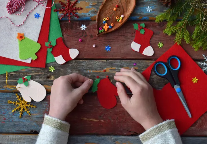 Manos creando adornos navideños con fieltro rojo y verde. Tijeras, cuentas y piñas completan la escena de manualidades.