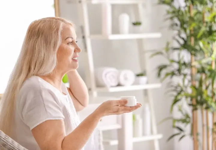 Uśmiechnięta kobieta z blond włosami trzyma słoiczek kremu, zastanawiając się, co jest dobre na cerę. W tle łazienka z roślinami.