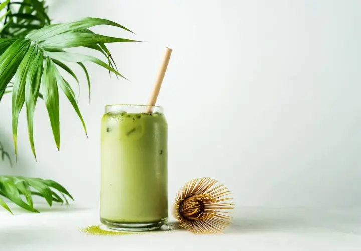 Orzeźwiające matcha latte z lodem i bambusową słomką, obok bambusowa miotełka. Poznaj niezwykłe matcha właściwości!