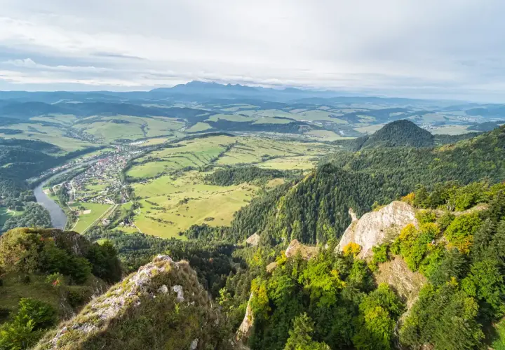Widok z góry na dolinę z rzeką i miasteczkiem, w oddali majaczą Tatry. Widać skaliste zbocza porośnięte lasem, a w tle trzy korony nazwy szczytów.