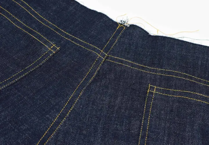 Close-up of the yoke on dark blue jeans, showing yellow stitching and raw edges.