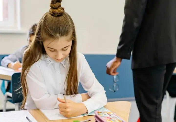 Dziewczynka skupiona na zadaniu podczas testu trzecioklasisty. Nauczyciel stoi obok, trzymając okulary.