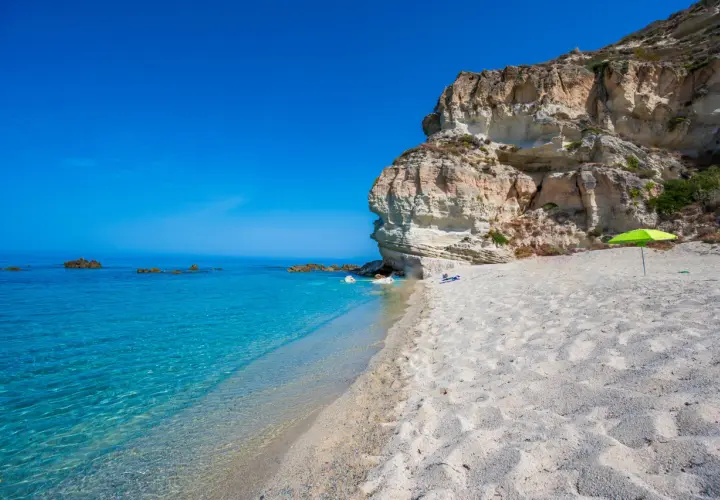 A secluded beach in Calabria, Italy, with crystal-clear turquoise water, white sand, and dramatic cliffs. A bright green umbrella offers shade on this idyllic spot.