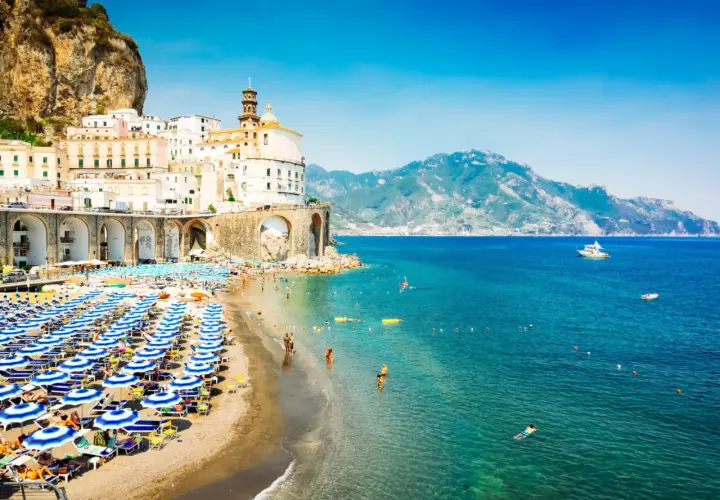 A vibrant beach scene in Naples, with rows of blue umbrellas and sunbathers. Buildings cling to a cliffside, overlooking the sparkling turquoise sea.