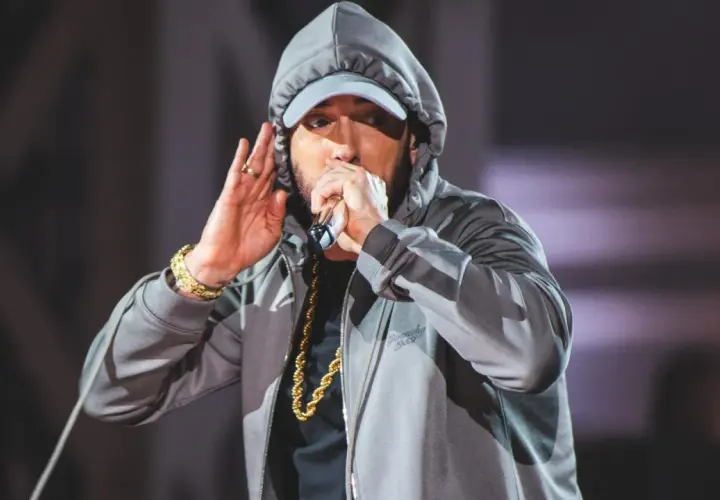 Eminem w szarej bluzie z kapturem i czapce, trzymający mikrofon i gestykulujący.
