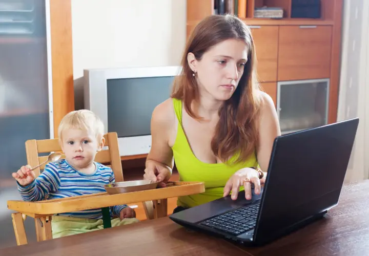 Mama na zasiłku macierzyńskim pracuje przy laptopie, podczas gdy jej dziecko siedzi w krzesełku i trzyma łyżeczkę.