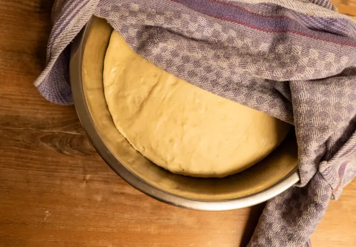 Ein goldgelber Pizzateig ruht in einer Schüssel, bedeckt mit einem karierten Tuch. Seine weiche, elastische pizzateig konsistenz verspricht eine köstliche Pizza.