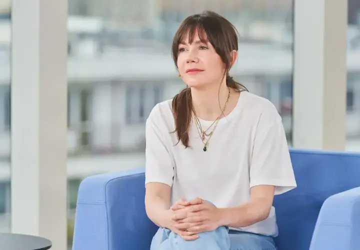 Olga Bołądź, której wiek dodaje jej uroku, siedzi w białej koszulce i jeansach, z dłońmi złożonymi na kolanach.