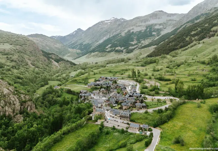 Pueblo de casas de piedra en un valle verde del Val d'Aran, rodeado de montañas cubiertas de vegetación y algunas cumbres nevadas.