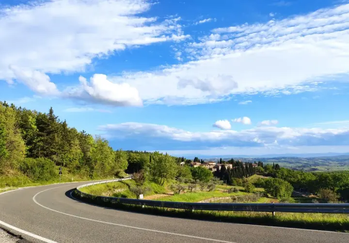 Strada SR 222 Chiantigiana serpeggia tra colline verdi e boschi, con un borgo toscano in lontananza sotto un cielo azzurro e nuvoloso.