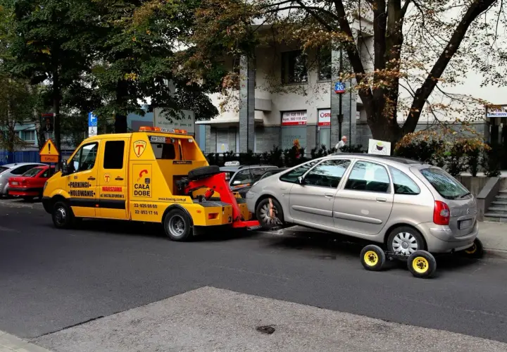 Żółty holownik zabiera srebrny samochód, który zapewne otrzymał mandat za parkowanie.