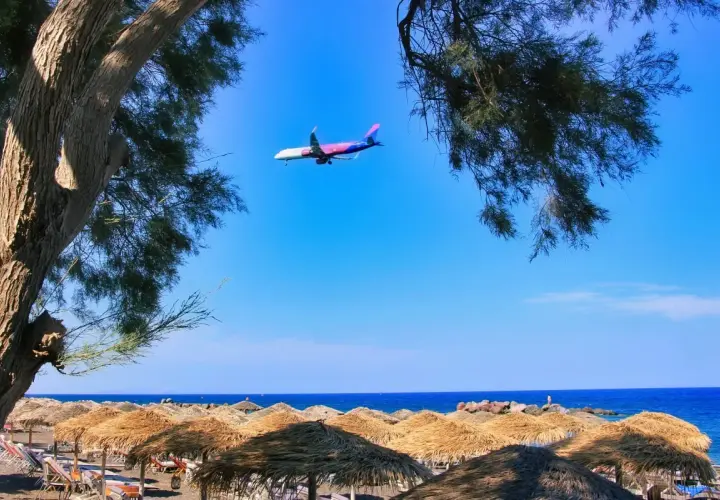 Samolot przelatuje nad plażą z parasolami, tuż przed lądowaniem na **Santorini lotnisko**.