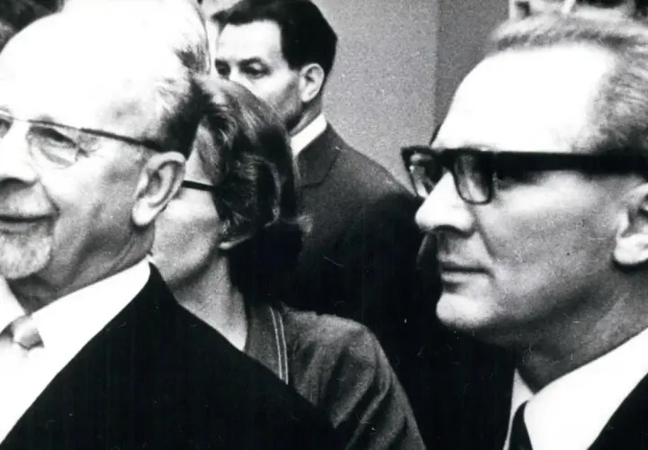 Ein Mann mit Brille und Bart, ein DDR-Chef, lächelt. Im Hintergrund weitere Personen.