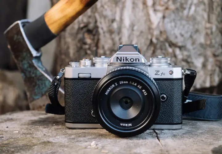 Srebrny aparat Nikon Zfc z obiektywem Nikkor Z 28mm f/2.8 SE na drewnianym tle. Obok siekiera. Test aparatu.