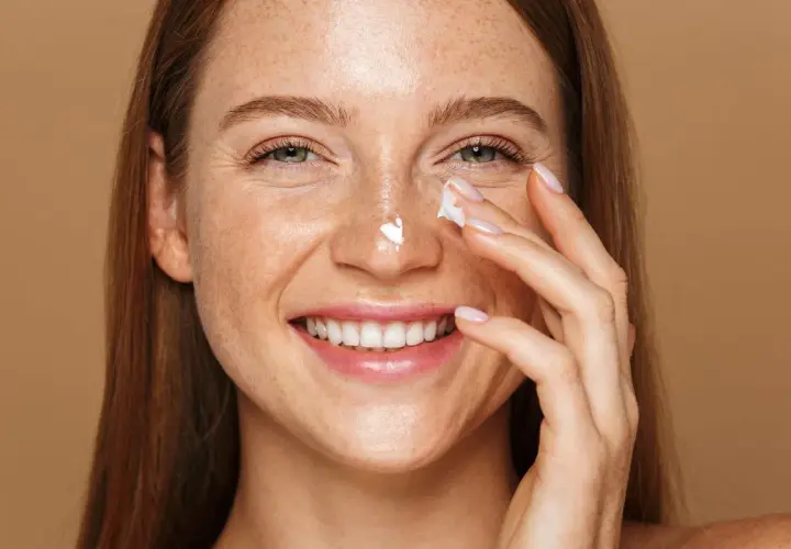Uśmiechnięta kobieta z piegami aplikuje krem na nos. Pamiętaj o ochronie przed słońcem, zwłaszcza stosując retinol.