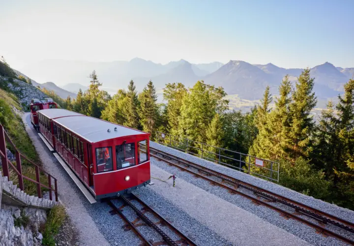 Czerwony wagon kolejki zębatej jedzie po torach w górę zbocza. W tle góry i lasy.