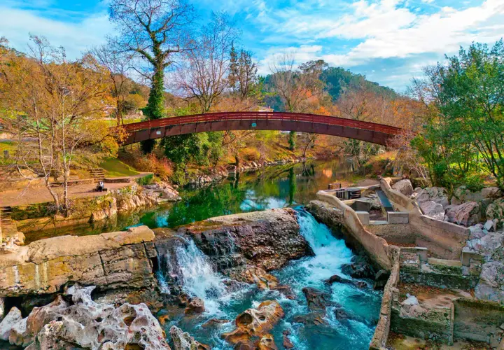 Cascada y puente de madera en la vía verde Puente Viesgo, rodeados de árboles otoñales y un cielo azul vibrante.