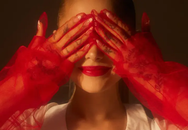 Ariana Grande 2024 w czerwonych rękawiczkach zakrywa oczy, odsłaniając usta i tatuaże na dłoniach.