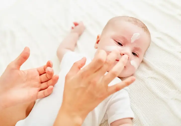Delikatna pielęgnacja noworodka. Krem na buzi maluszka, troskliwe dłonie mamy.