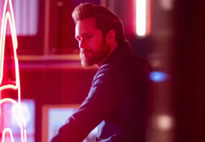 Mężczyzna z brodą w ciemnej koszuli odwraca się od neonowego napisu. Scena z programu telewizyjnego "Bastion".