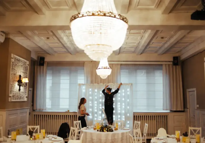 Dwie kobiety przygotowują salę weselną, jedna stoi na drabinie, dekorując tło światełkami. To początek drogi, jak zostać wedding plannerką.