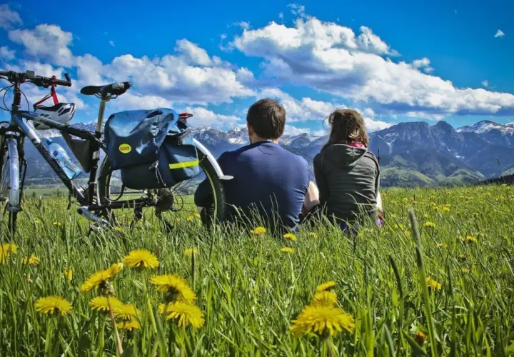 Para na rowerach odpoczywa na łące pełnej mleczy, podziwiając Tatry. To idealne miejsce na szlak papieski Gubałówka.