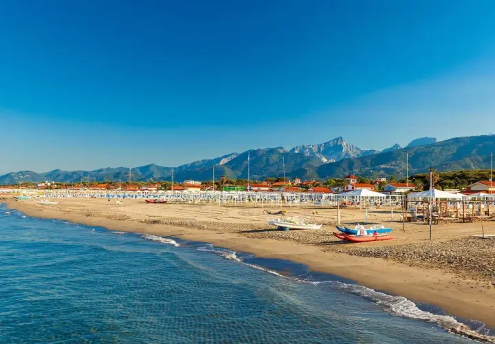 Słoneczna plaża w Toskanii, z rzędami białych parasoli i leżaków, w tle góry i błękitne morze.
