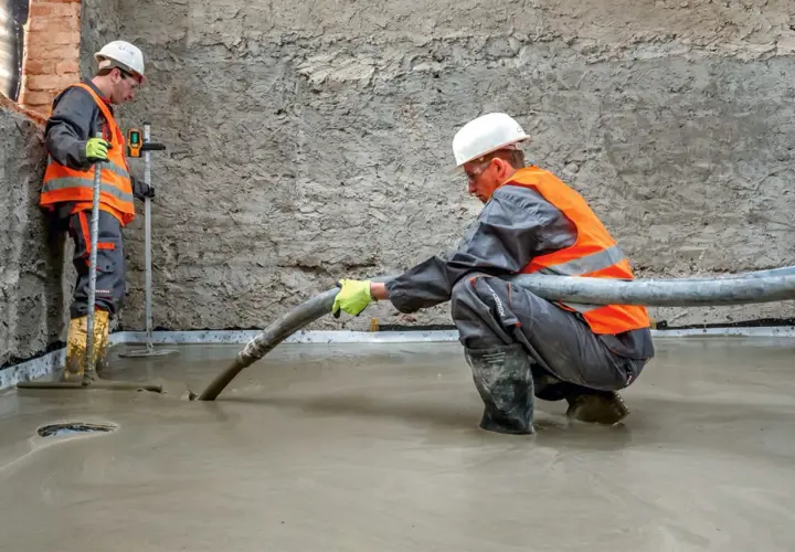 Dwóch budowlańców w kamizelkach odblaskowych wylewa beton. Jeden kieruje wąż z masą, drugi kontroluje poziom.