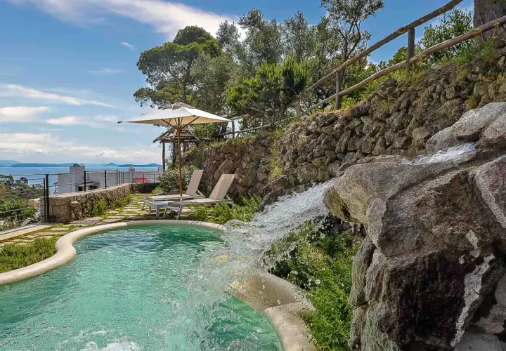 Piscina con cascata che ricorda le terme naturali di Ischia, circondata da vegetazione e con vista sul mare.