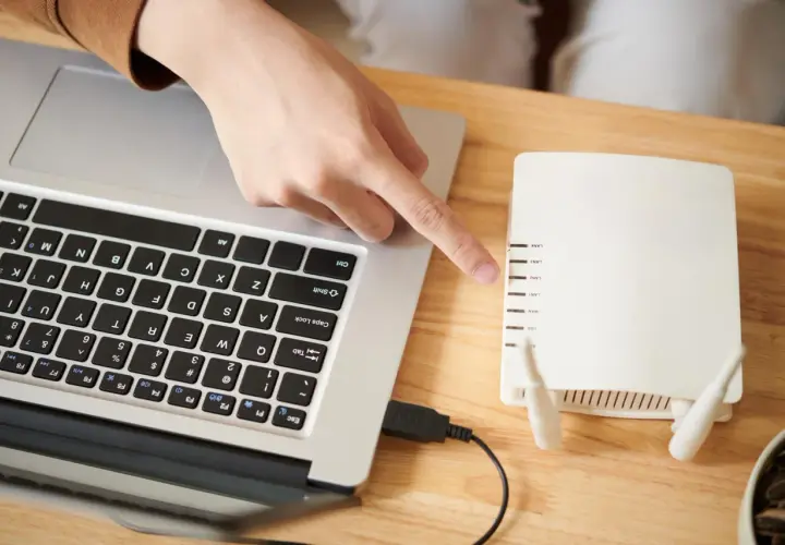 Dłoń wskazuje na router, podłączony kablem do laptopa. Dowiedz się, jak włączyć wifi w laptopie, korzystając z tego połączenia.