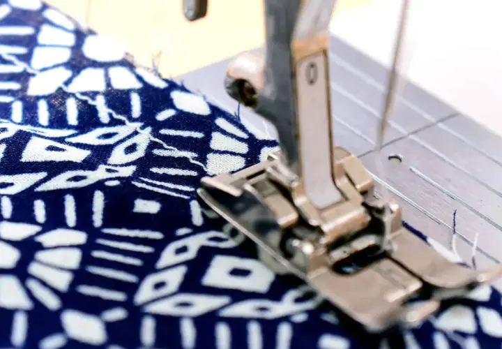 Sewing machine foot creating a narrow hem on a blue and white patterned fabric.