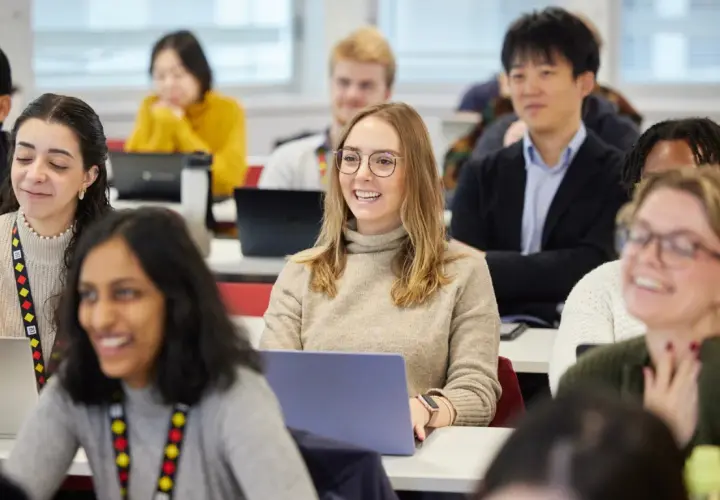 Students in a classroom, some with laptops, are engaged in a lesson. One student smiles, perhaps anticipating her MPA credential.