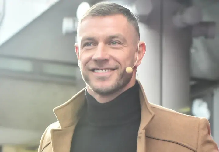Uśmiechnięty Dariusz Pachut w beżowym płaszczu i czarnym golfie, z mikrofonem przy uchu. Może opowiada o swojej żonie.