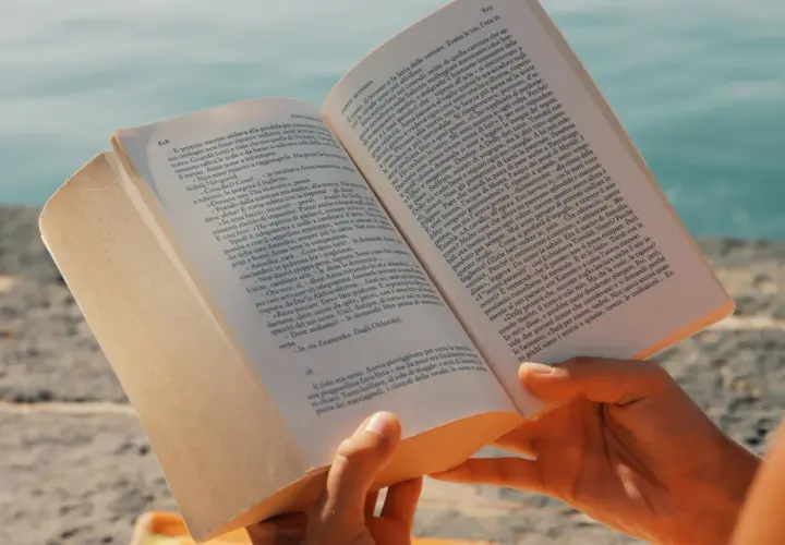 Mani che sfogliano un libro aperto, con un mare azzurro sullo sfondo. Un'immagine che evoca la lettura di una **canzone in dialetto siciliano** sotto il sole.