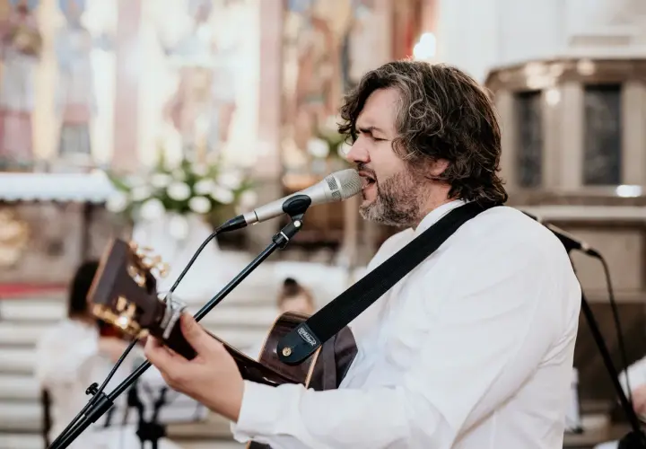 Mężczyzna z gitarą śpiewa pieśni chrześcijańskie w kościele. W tle widać ołtarz i obrazy.
