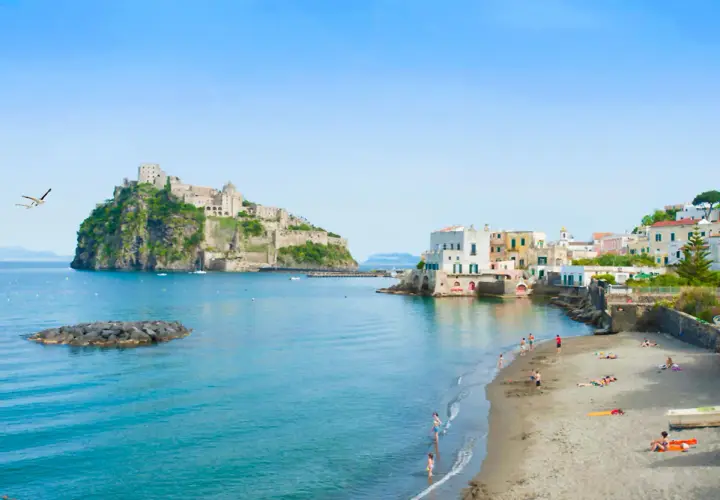 Lidi di Ischia Porto con il Castello Aragonese sullo sfondo. Gente in spiaggia e mare azzurro sotto un cielo limpido.