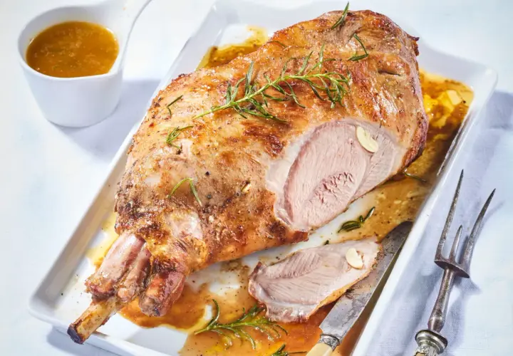 Gigot d'agneau désossé au four, doré et juteux, garni de romarin et d'ail. Servi avec sa sauce dans un plat blanc.