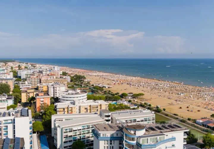 Widok z lotu ptaka na zatłoczoną bibione plażę, pełną białych parasoli i ludzi. W tle błękitne morze i nadmorskie miasteczko.